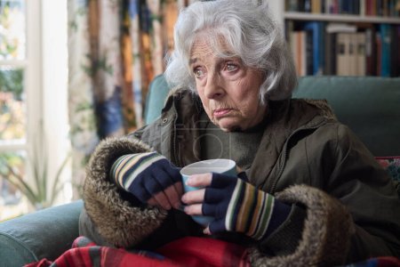 Foto de Mujer mayor usando abrigo en interiores con bebida caliente tratando de mantener caliente en casa en crisis de energía - Imagen libre de derechos
