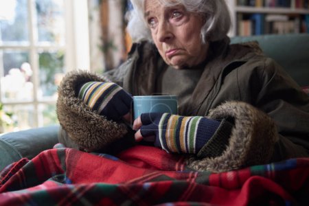 Foto de Primer plano de la mujer mayor que usa abrigo en interiores con bebida caliente tratando de mantener caliente en casa en la crisis energética - Imagen libre de derechos