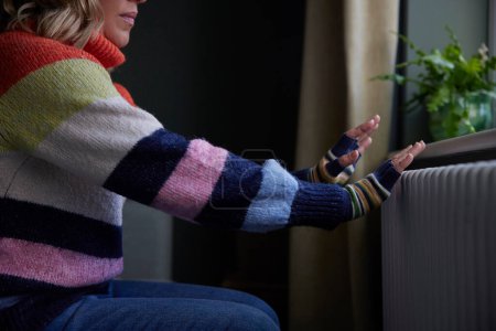 Mujer en casa en guantes que calientan las manos por el radiador durante el costo de la crisis de energía de vida