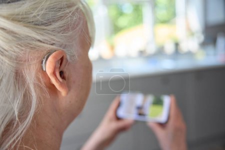 Foto de Primer plano de la mujer mayor que lleva detrás del audífono del oído o ayuda Sreaming desde el teléfono móvil en casa - Imagen libre de derechos