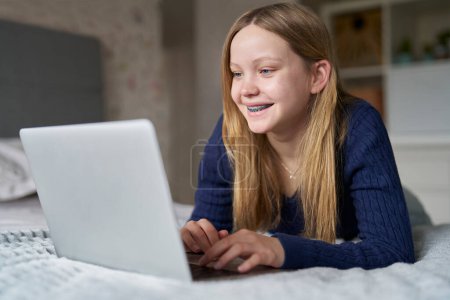 Adolescente chica usando ortodoncia aparatos ortodónticos con el ordenador portátil acostado en la cama en casa Juegos, Streaming Film or Show, Navegación en línea Mirando a las redes sociales