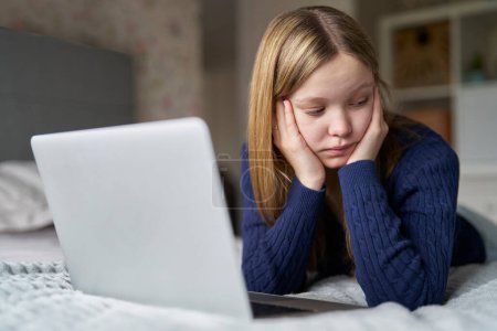 Niña adolescente infeliz con computadora portátil acostada en la cama en casa ansiosa por el acoso en línea en las redes sociales y estar en línea demasiado 