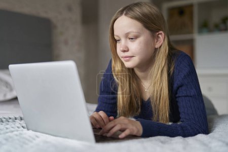 Chica adolescente con computadora portátil acostada en la cama en casa Juegos, Streaming Film or Show, Navegación en línea Mirando a las redes sociales