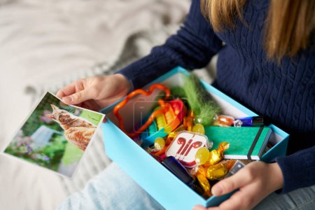 Close Up Of Teenage Girl Sitting On Bed At Home Using Soothe Box Containing Pet Photo, Fidget Toy, Candy Or Sweets, Earphones For Music, Fabric And Scented Spray To Help With Stress And Anxiety