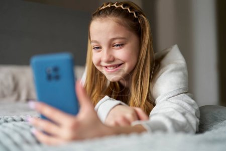 Sonriente adolescente con uñas pintadas acostada en la cama en casa usando teléfono móvil