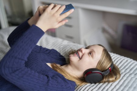  Teenagermädchen mit Zahnspangen liegen zu Hause auf dem Bett und streamen Musikfilme oder zeigen vom Telefon auf drahtlose Kopfhörer