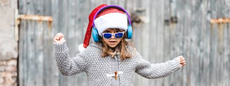 Photo for Horizontal banner or header with funny little girl wearing Santa Claus hat and sunglasses dancing against a wooden background outdoors in Christmas time - Childhood concept in Christmas days - Royalty Free Image