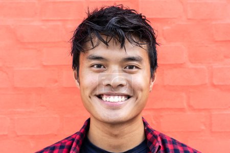 Foto de Retrato de joven asiático guapo de pie al aire libre contra un fondo de pared roja en la ciudad y mirando a la cámara - Retrato chico asiático feliz - Generación z concepto de la gente - Imagen libre de derechos