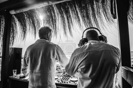 Téléchargez les photos : Back view of disc jockeys playing music for tourist people at club party outdoors on the beach - Djs wearing headphones at music live event - Music and fun concept - Black and white editing - en image libre de droit