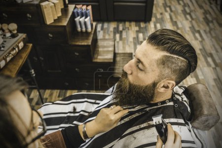 Foto de Hipster man at barbershop salon getting beard and hair cut - Hairdresser woman using hair clipper and comb for to modern gentleman cut - Barber shop concept - Imagen libre de derechos
