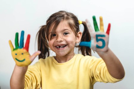 Foto de Happy toothless little girl with the five number painted on the hand laughing and having fun - Little girl painting her hands with smiley faces and numbers - The number five and childhood concept - Imagen libre de derechos