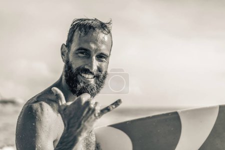 Surfer happy with surf surfing smiling doing hawaiian Shaka Brah or Hang Loose during surf session in ocean waves on beach vacation -  Friendly greeting in surfer culture - Black and white editing