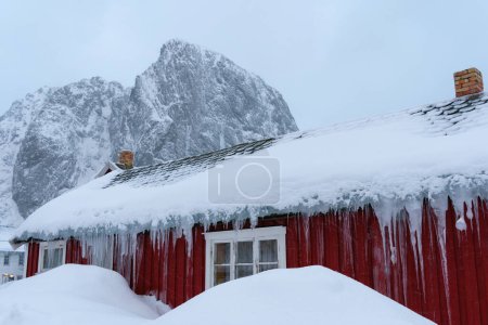 Foto de Montaña de piedra en forma de corazón detrás de la famosa atracción turística Hamnoy pueblo de pescadores en las Islas Lofoten, Noruega con casas rojas rorbu. Con nieve cayendo en invierno. - Imagen libre de derechos