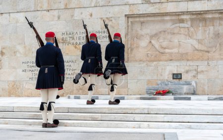 Photo for Text on the wall "And one bed remains empty for the unknown" in Greek and greek evzone soldiers in traditional costumes guard of honor at a monument in greece - Royalty Free Image