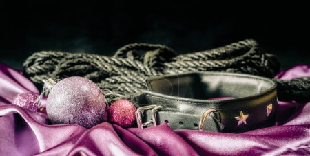 bdsm bodegón negro collar de cuero cuerda shibari y bola de Navidad lila en tela rosa
