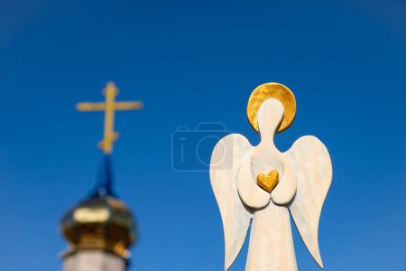 Foto de Figura de madera de un ángel con un halo dorado y el corazón en las manos sobre un fondo de la iglesia. Cielo azul. - Imagen libre de derechos