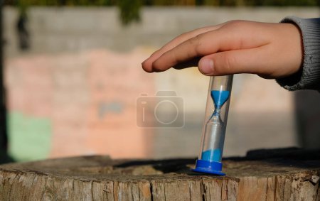 Photo for Child's hand with sandglass or sand timer in the home yard. Wall background. Sunlight. Copy space. - Royalty Free Image