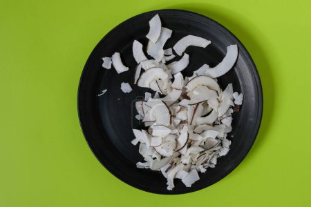 Dried Organic Coconut. Chips. Black ceramic plate. Yellow-green color background. Top view. 