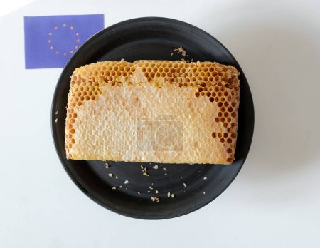 A piece of honeycomb on a plate. Sealed honeycombs. Unfilled edges. Euro Union flag. Export or import honey.