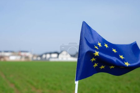 Flagge der Europäischen Union. Weizenfeld im Frühling. Unscharfer Hintergrund eines wohlhabenden Dorfes. Kopierraum.