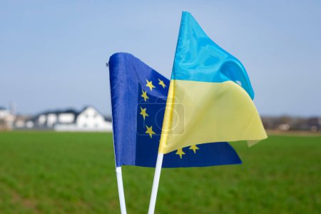 Flagge der Europäischen Union. Ukrainische Flagge. Weizenfeld im Frühling. Unscharfer Hintergrund eines wohlhabenden Dorfes. Die Ukraine hat den Status eines Beitrittskandidaten zur Europäischen Union.