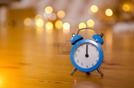 Blue small arrow alarm clock on the wooden floor against the background of the Christmas tree, bed, garlands and yellow bokeh from them, on the clock it is midnight, the time of the new year