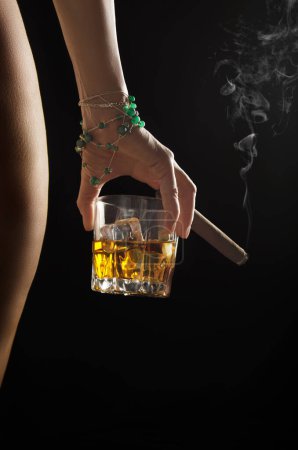 Photo for Caucasian model holds glass of whiskey with icecubes and smoking cigar in her hand near naked buttock and hip on dark background closeup studio shot - Royalty Free Image