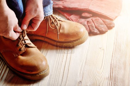 Foto de Carpintero en jeans azules atando cordones de botas de trabajo amarillas sobre suelo de madera. Lugar para el texto - Imagen libre de derechos