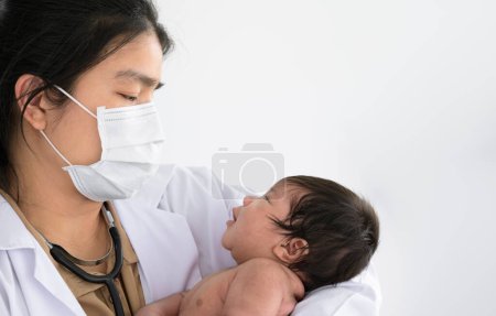 Female pediatrician doctor with face mask holding happy asian newborn baby girl or boy in her arms for general physical check up  in clinic. Baby healthcare concept.