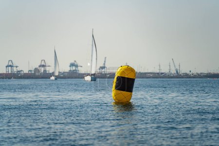Téléchargez les photos : Yellow buoy, sailboat competition in the background with commercial port - en image libre de droit