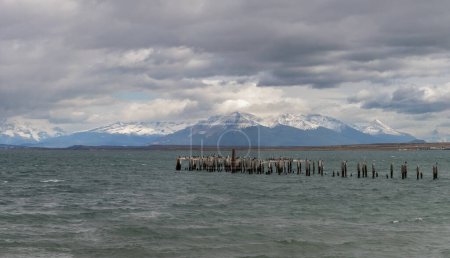 Wooden posts in choppy sea before snowy mountains.
