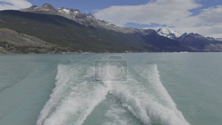 Slow-mo footage of a yacht making waves on a glacier lake against a backdrop of lush green and blue mountains.