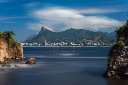 Impresionante vista del mar golpeando rocas en una costa, con la estatua icónica de Cristo Redentor de Río de Janeiros bajo nubes dramáticas.