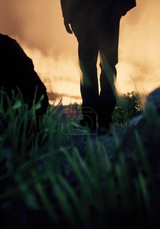 Foto de El hombre de traje negro camina entre la hierba alta en un paisaje rocoso durante la puesta del sol. Vista trasera. Renderizado 3D. - Imagen libre de derechos