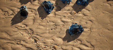 Téléchargez les photos : Des liasses de papier bleu sale recouvert d'huile noire gisant dans le sable ondulé de la plage. - en image libre de droit