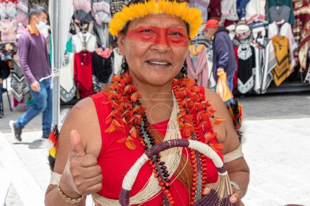 Foto de Cuenca, Ecuador-25 de marzo de 2023: Feria Artesanal de la Provincia de Pastaza. Mujer indígena de la etnia Huaorani (Waorani) en vestimenta típica y pintura facial presenta una artesanía hecha de fibra de palma. - Imagen libre de derechos