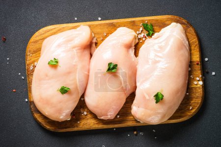 Téléchargez les photos : Poitrine de poulet avec épices à la planche à découper en bois sur table noire. Vue du dessus. - en image libre de droit