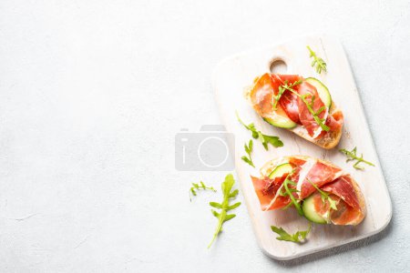 Foto de Open sandwiches with cream cheese, prosciutto and arugula at white background. Top view with copy space. - Imagen libre de derechos