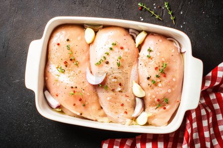 Téléchargez les photos : Poitrine de poulet dans le plat de cuisson avec des épices. Viande crue prête à cuire. Vue de dessus à la table de cuisine sombre. - en image libre de droit