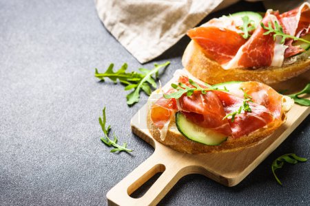 Photo for Appetizer crostini, open sandwiches with cream cheese, prosciutto, cucumber and arugula at cutting board. - Royalty Free Image