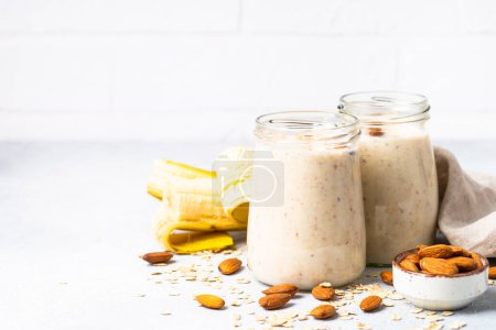 Photo for Almond banana smoothie with oat flakes in glass jars at white table. - Royalty Free Image