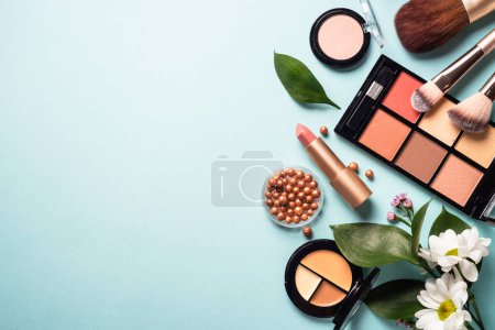 Foto de Cosmetic products on blue background. Cream, powder, shadow, brushes with green leaves and flowers. Flat lay image with copy space. - Imagen libre de derechos