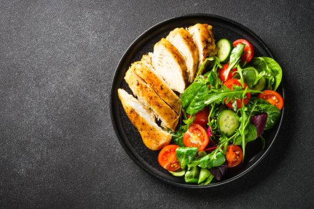 Foto de Healthy food plate, keto diet. Green salad with chicken fillet on black table. Top view with copy space. - Imagen libre de derechos