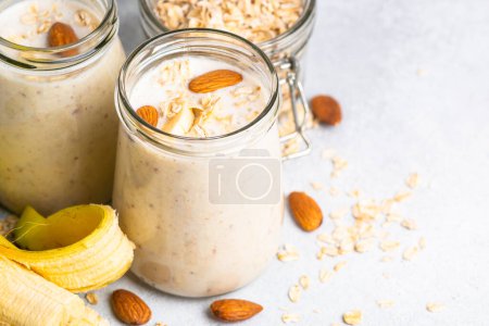 Téléchargez les photos : Almond banana smoothie with oat flakes in glass jars at white table. - en image libre de droit