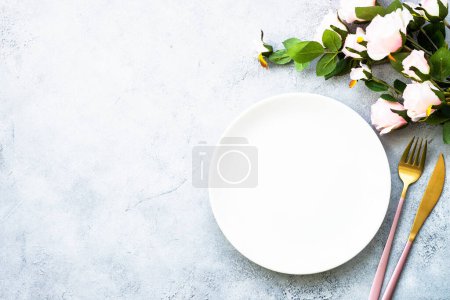 Foto de Ajuste de mesa con plato blanco, cubiertos modernos y rosas rosadas. Piso en blanco con espacio de copia. - Imagen libre de derechos