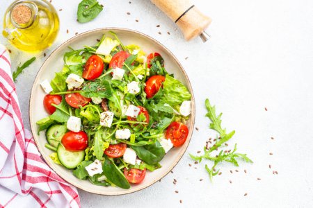 Téléchargez les photos : Salade de printemps dans une assiette artisanale. Épinards, roquette, tomates et feta à l'huile d'olive. Vue de dessus sur blanc. - en image libre de droit