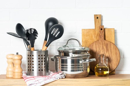 Kitchen table with kitchen utensils, cooking pots, oil bottle with wooden cutting board, white modern interior.