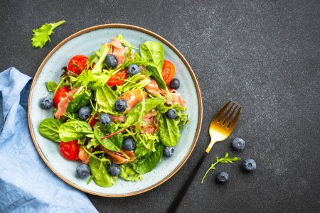 Téléchargez les photos : Salade verte au prosciutto frais, feuilles vertes mélangées et tomates à table noire. Vue supérieure avec espace de copie. - en image libre de droit