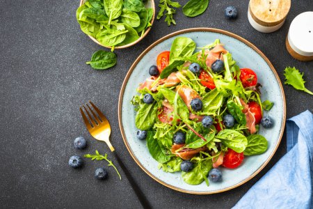 Photo for Healthy salad with prosciutto, green leaves mix and tomatoes at dark background. Top view with copy space. - Royalty Free Image