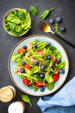 Photo for Green salad with fresh prosciutto, green leaves mix and tomatoes at dark table. Flat lay, vertical image. - Royalty Free Image